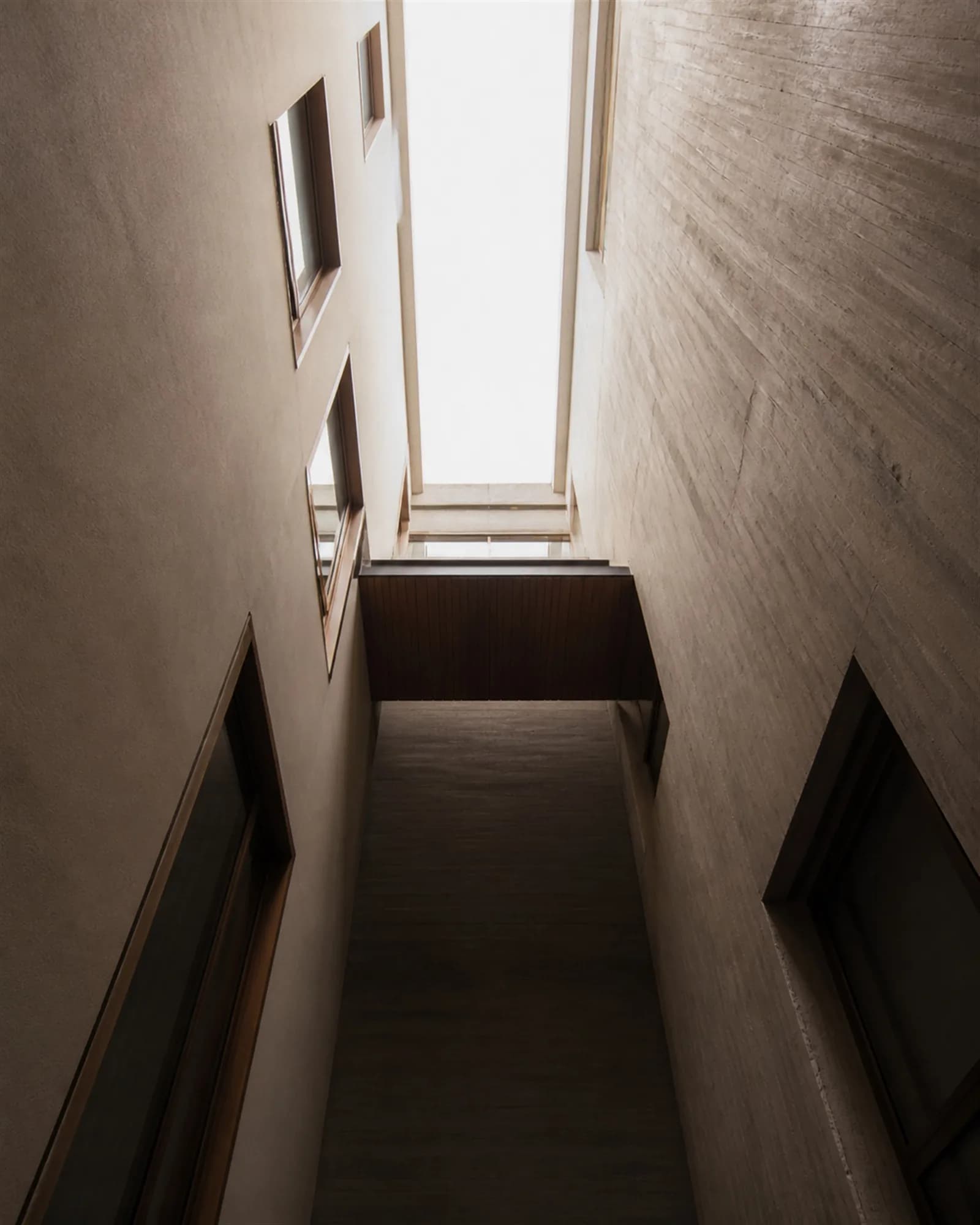 Looking up a three-storey internal light slot, walls receding toward bright open sky with a teak walkway crossing the void