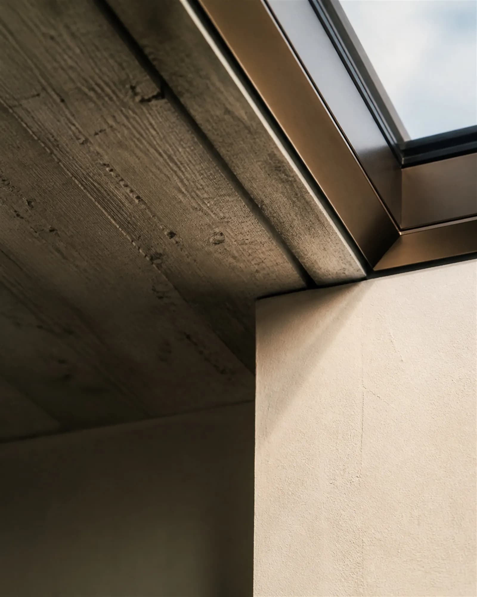 Close detail of a skylight junction where board-formed concrete meets a bronze frame and plaster reveal