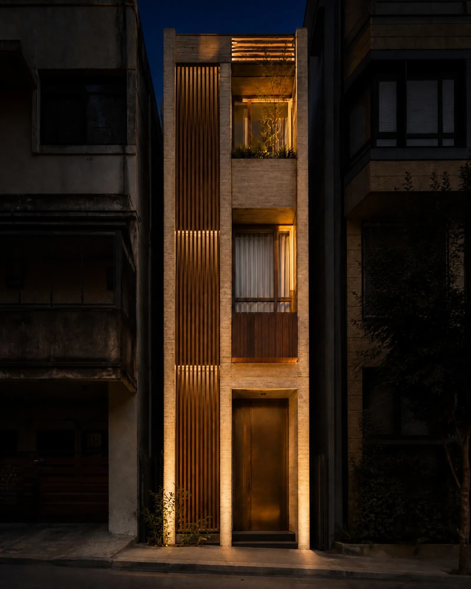 The narrow facade at night, travertine grazed by uplighting and the teak screen glowing from behind