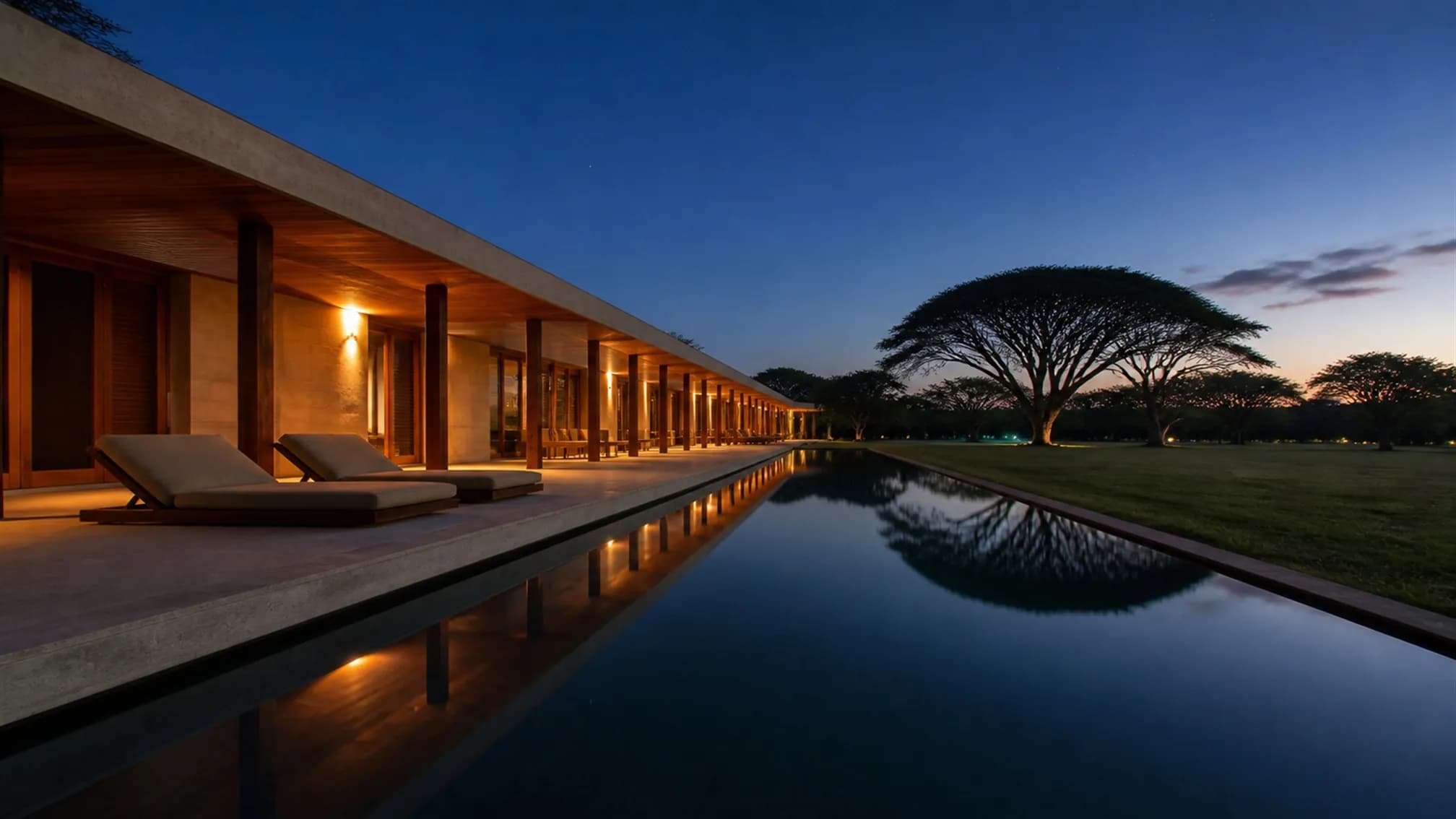 A long narrow lap pool at dusk running parallel to the house, reflecting the warmly lit verandah