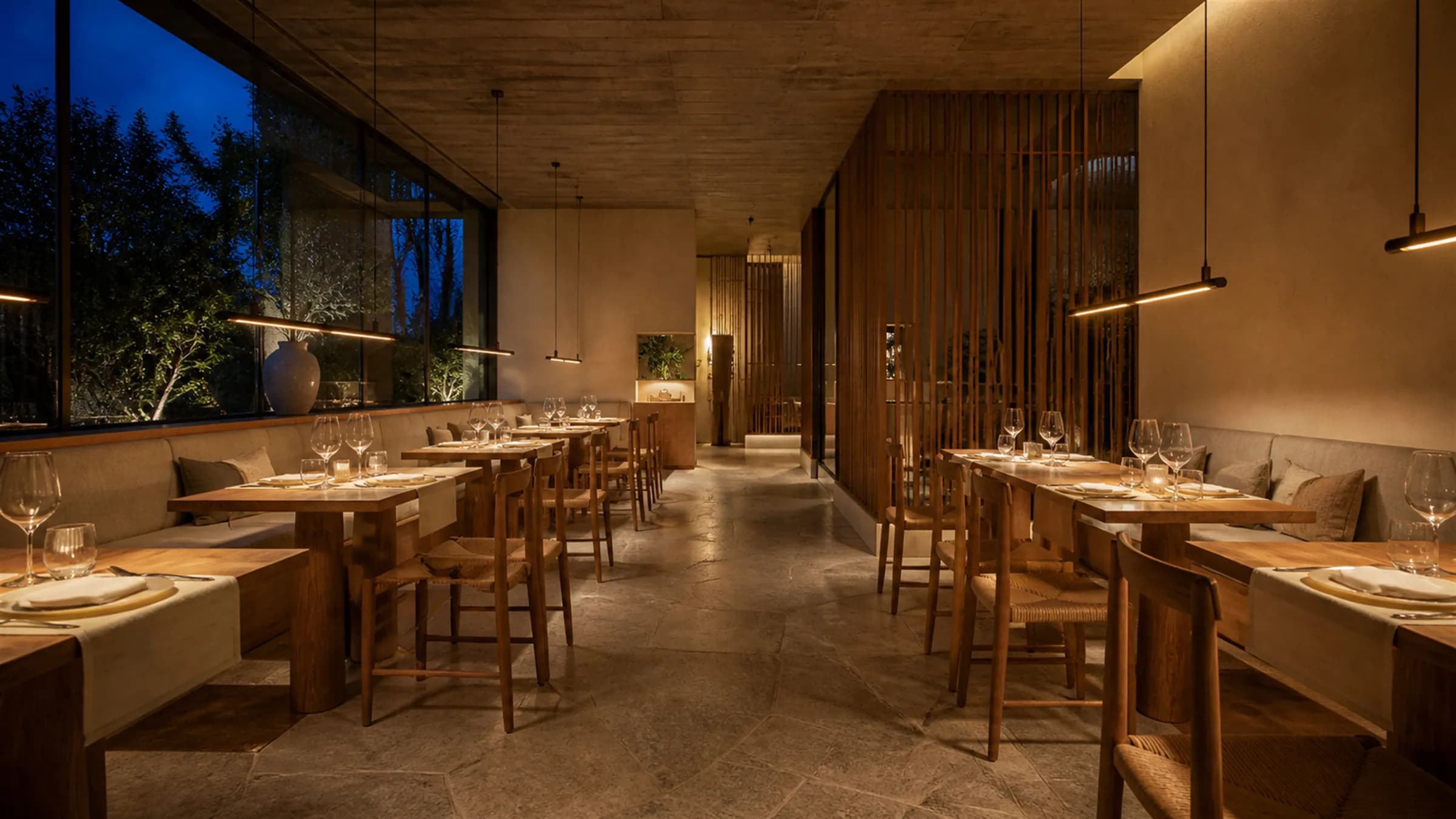 A restaurant interior at dusk, set and empty before service, with linear bronze pendants over teak tables